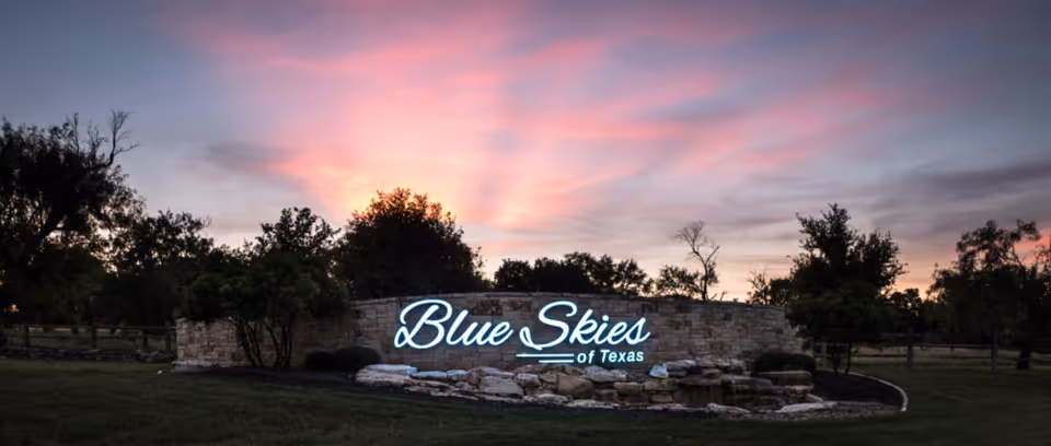 Stone entrance sign reading Blue Skies of Texas at sunset, framed by trees and lawn.