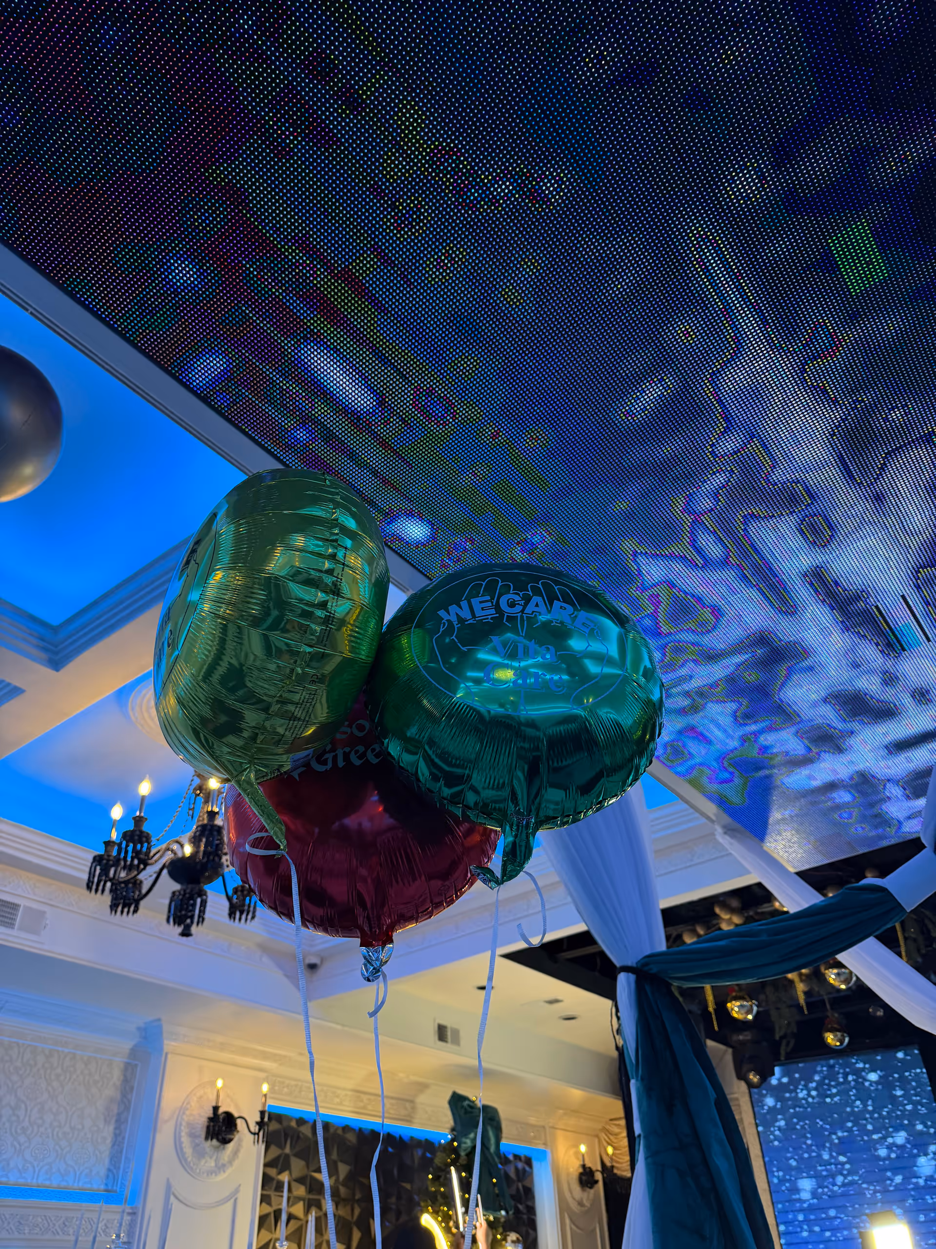 Indoor event space with teal balloons, white drapery, and ornate chandeliers.