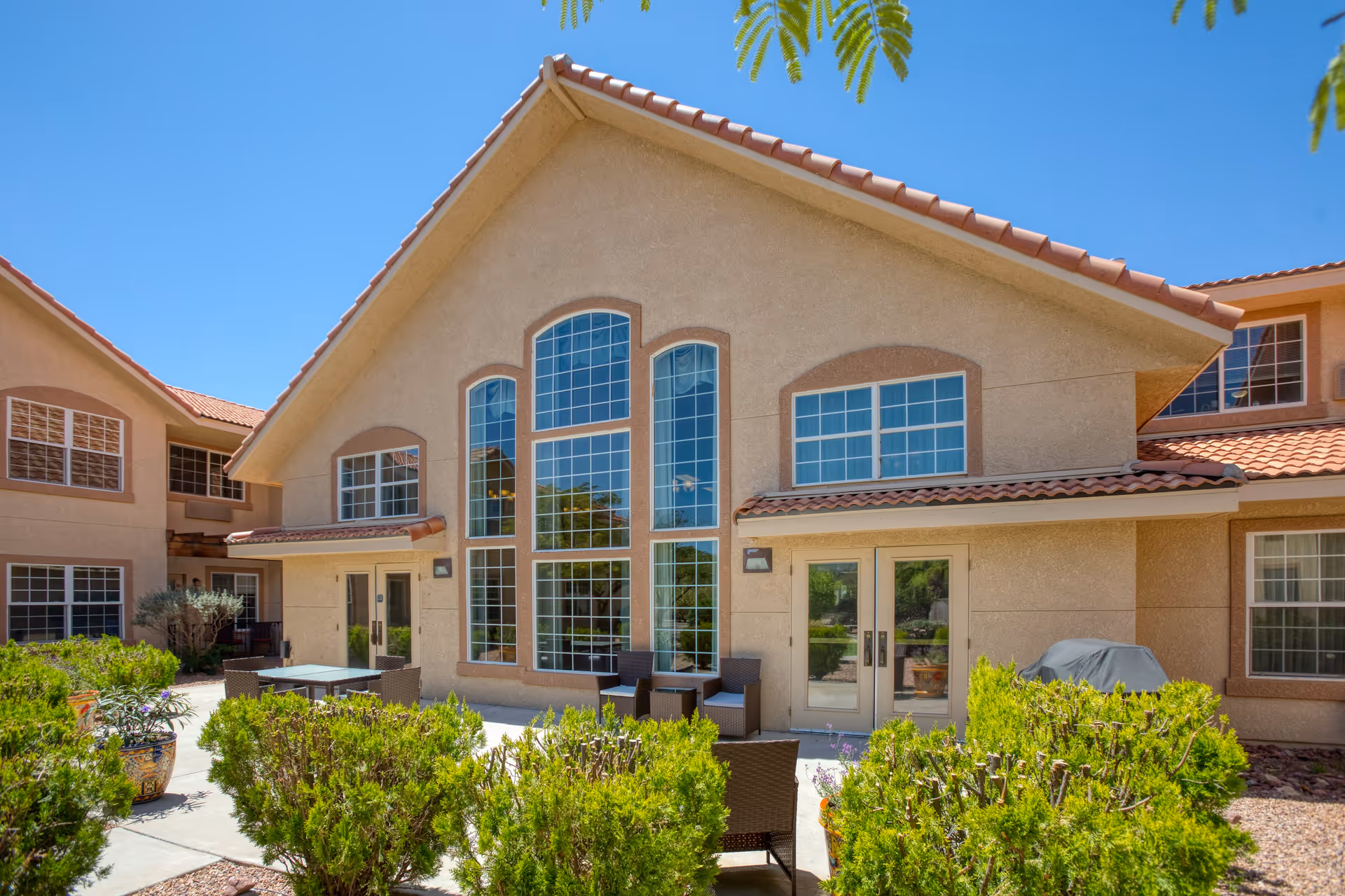 Exterior view of a senior living facility building with large windows and a patio area featuring outdoor seating and greenery under a clear blue sky.