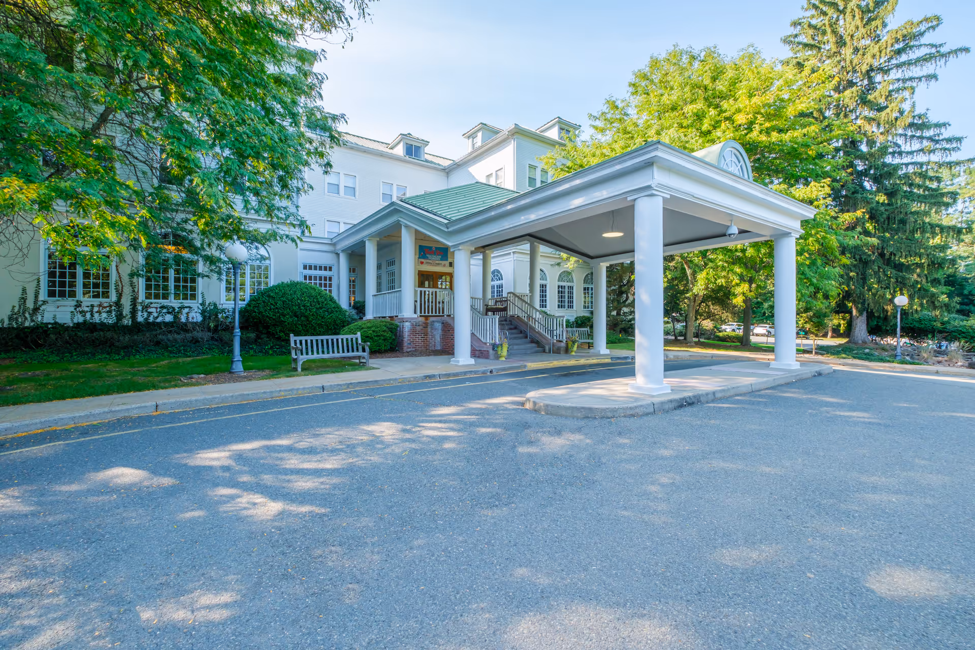 Front exterior view of a senior living facility named Green Hill Senior Living and Rehabilitation, featuring a white building with multiple windows, a green roofed entrance canopy supported by white columns, benches, and surrounding trees and greenery under a clear blue sky.