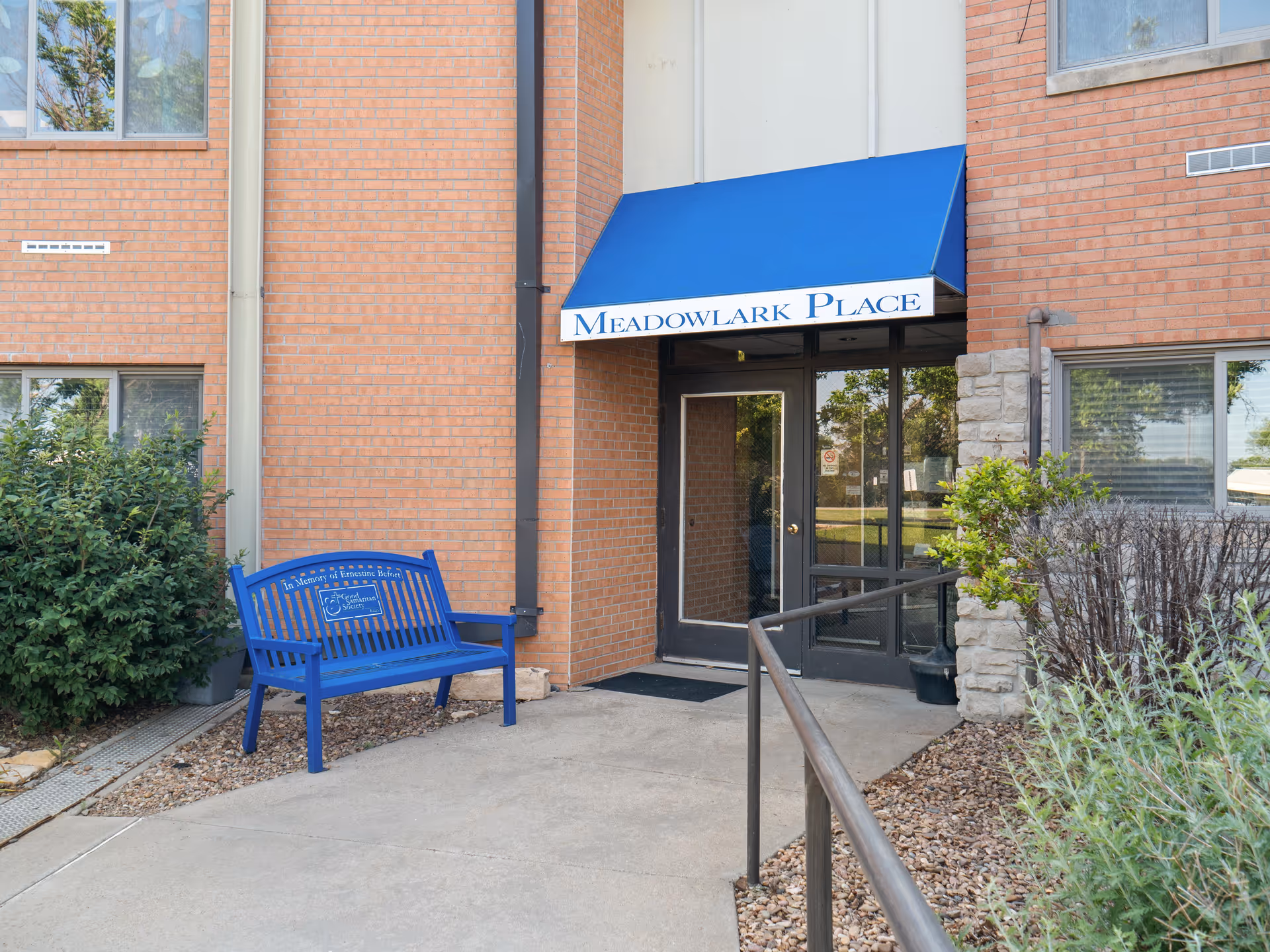 Brick building entrance with blue awning labeled Meadowlark Place and a blue bench.
