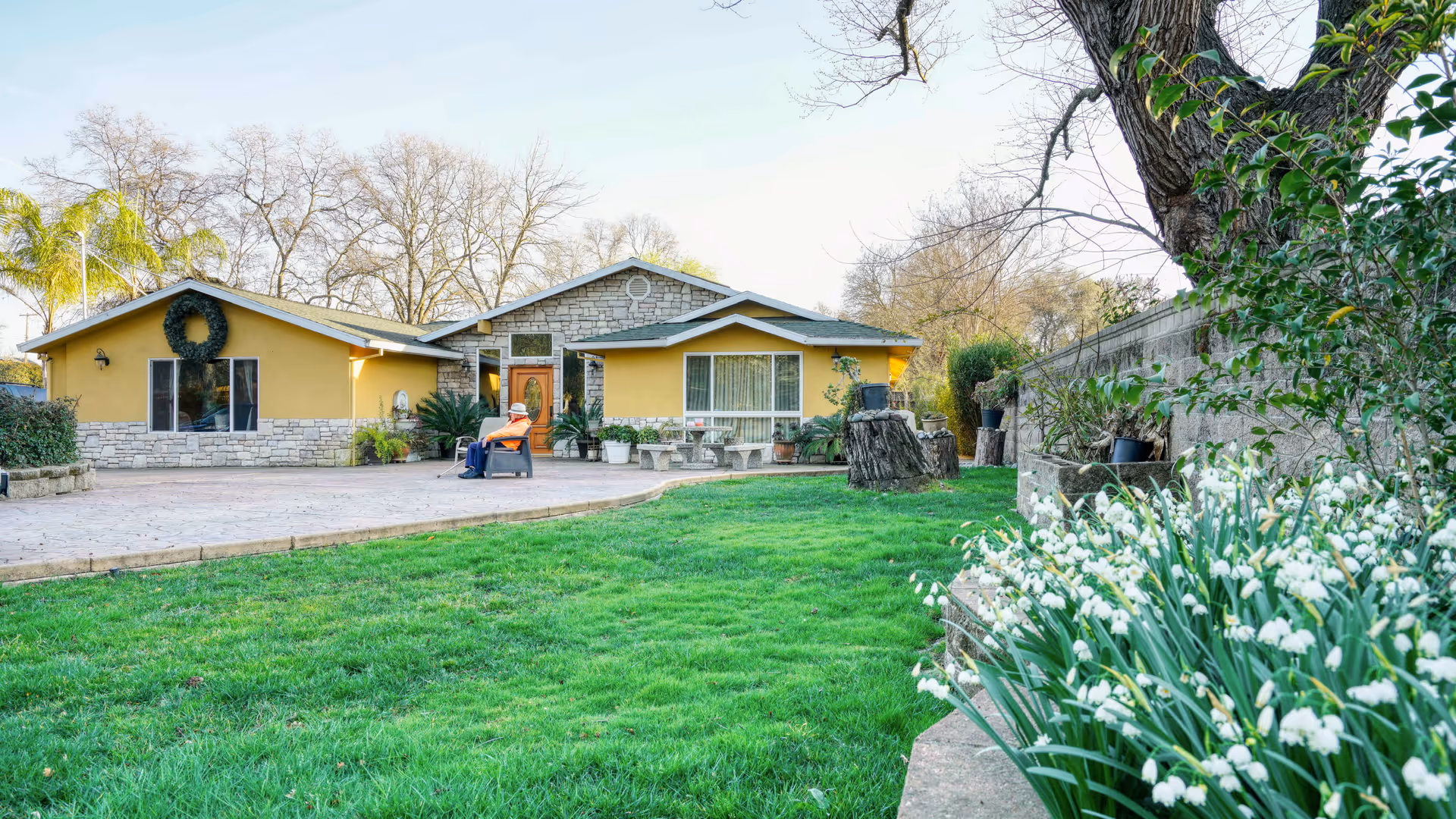 A single-story yellow building with stone accents, surrounded by a green lawn and trees. A person is sitting on a chair on the paved area in front of the building. White flowers and various plants are visible along the right side near a stone wall.
