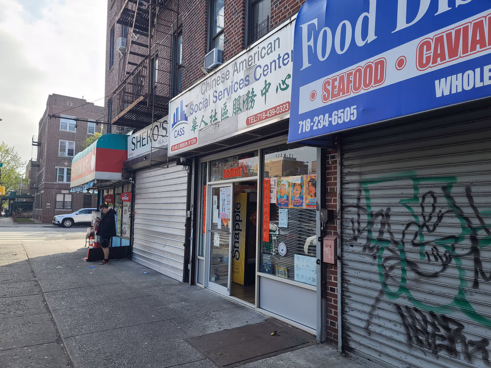 City street with storefronts, graffiti-covered shutter, and a person standing outside.