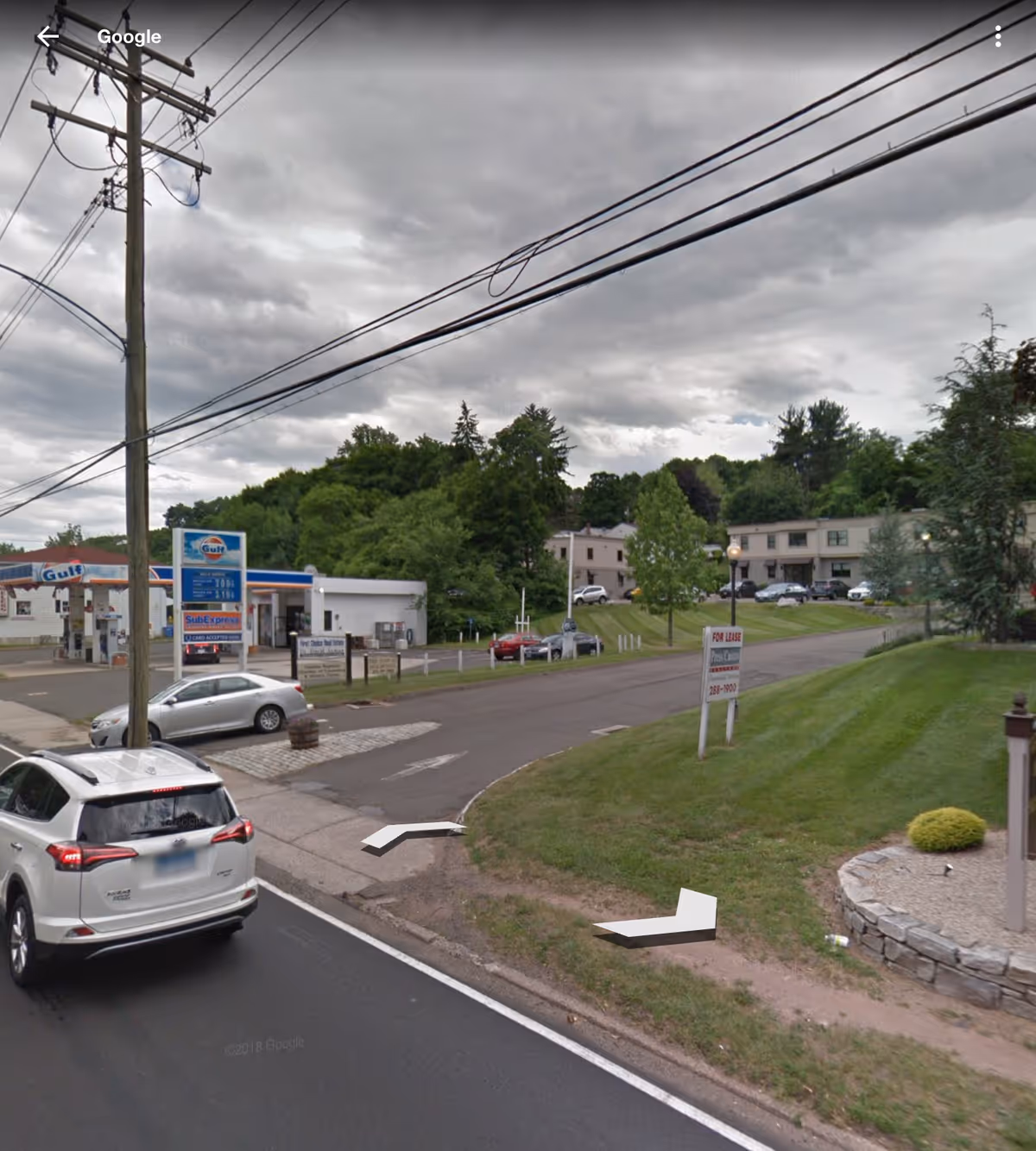 Wide street view showing a gas station, parked cars, and a grassy lot near residential buildings.