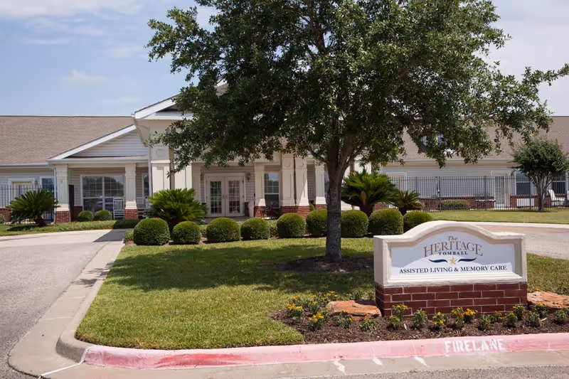 Front exterior of The Heritage Tomball Senior Living with landscaped shrubs and a sign.