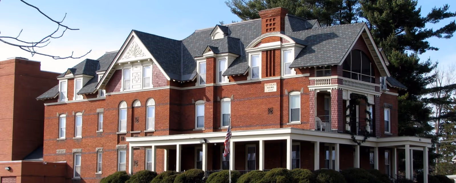 Front view of a large historic red-brick Victorian-style senior home with a wraparound porch, gabled roof, shrubs, and an American flag.
