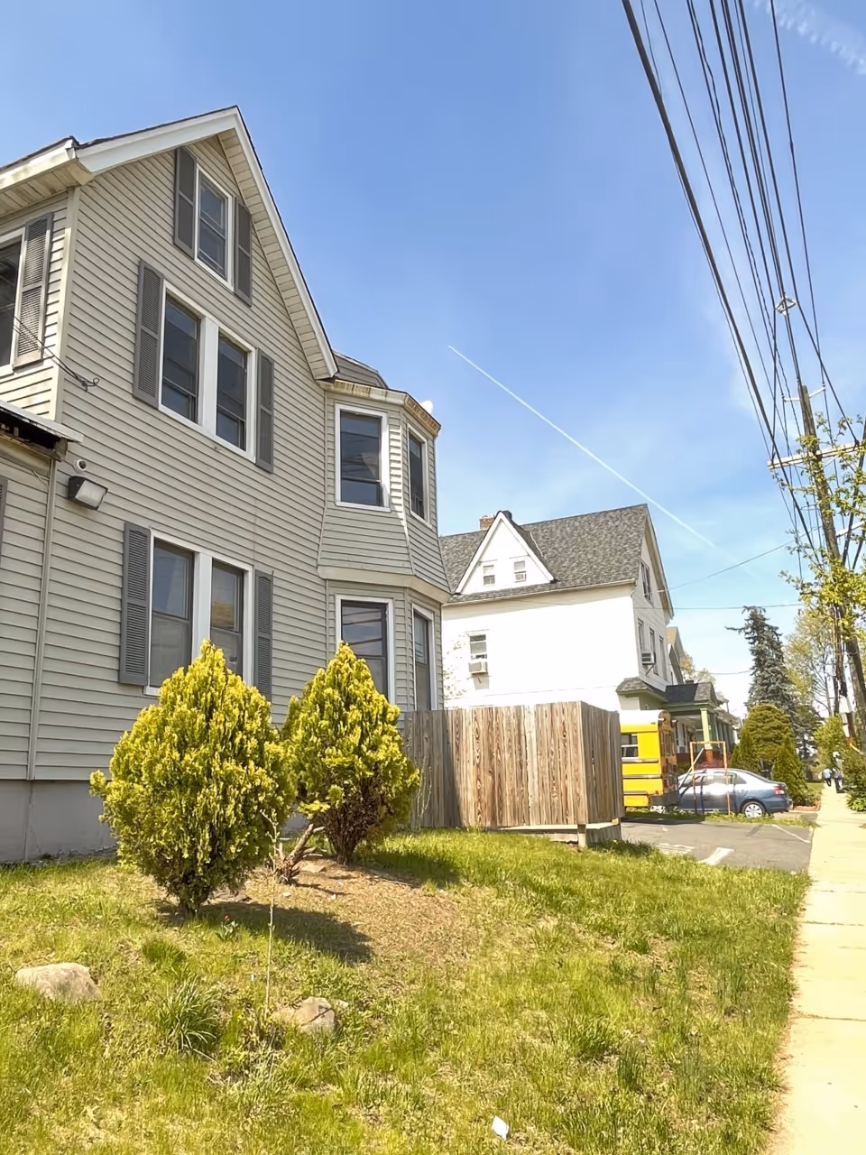 Exterior view of a multi-story house with siding, windows, greenery, fence, and power lines.