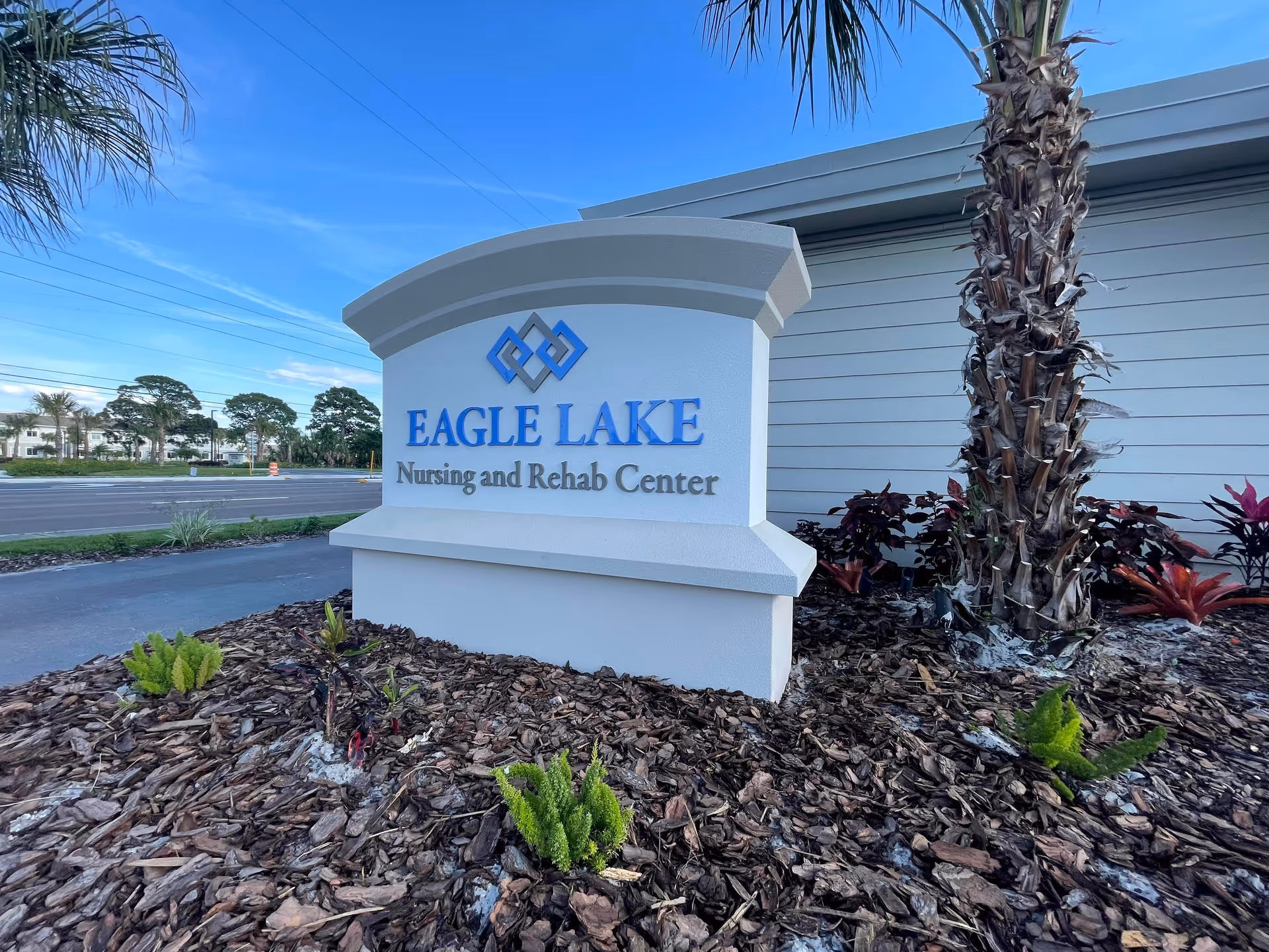 Outdoor view of a white sign with blue and gray lettering that reads 'Eagle Lake Nursing and Rehab Center' surrounded by landscaping with small plants and a palm tree, with a road and trees in the background under a clear blue sky.