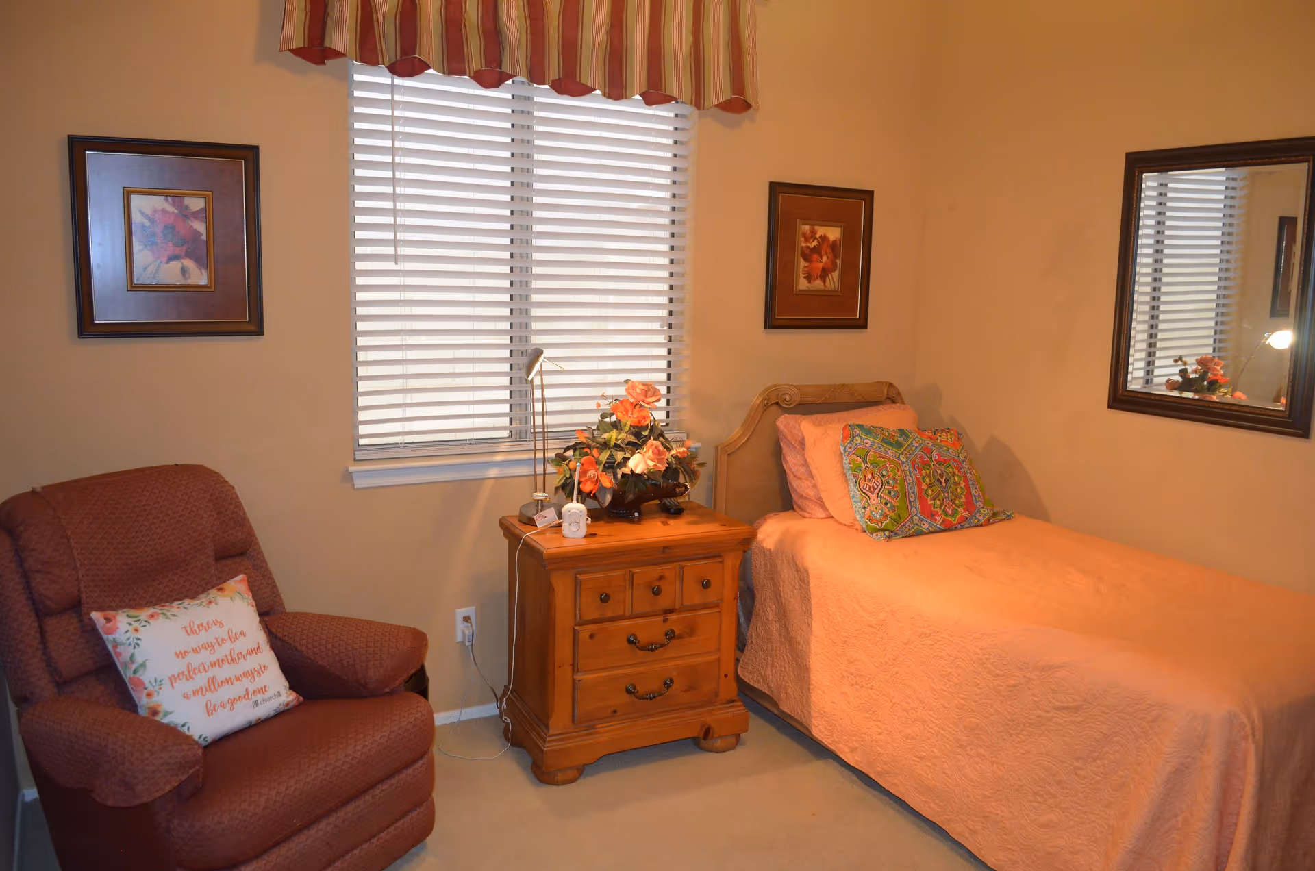 Cozy bedroom with a single bed covered in a pink quilt, a wooden nightstand, an upholstered armchair, window blinds and framed wall art.