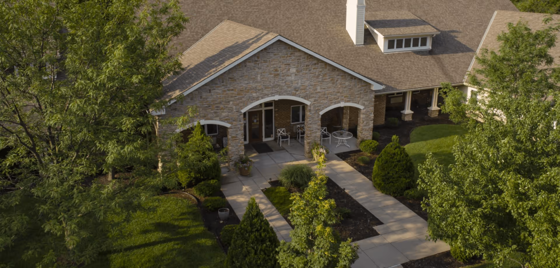 Stone-faced assisted living building front with arched entry, paved walkway, patio seating, and landscaped trees.