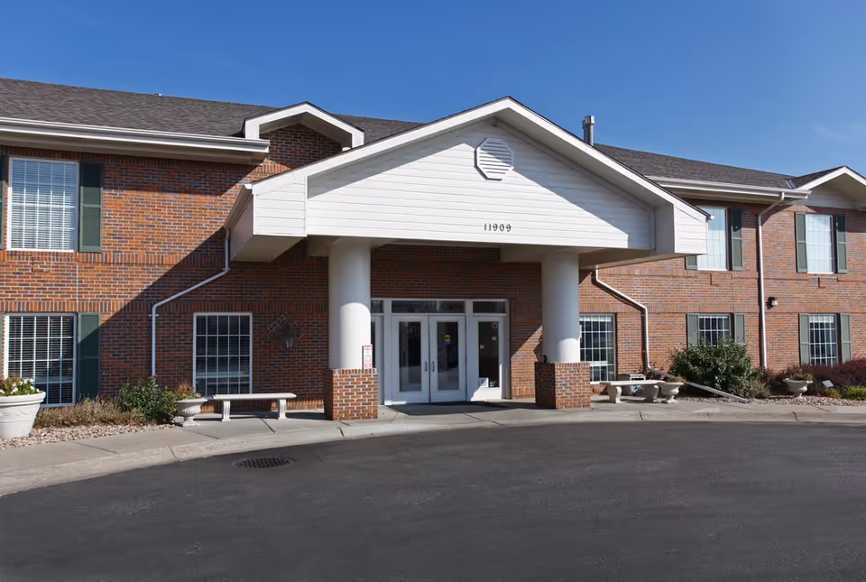 Brick two-story building façade with a white portico entrance, double glass doors, benches, and a driveway under a clear blue sky.