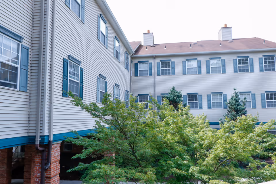 Exterior view of a multi-story assisted living facility building with beige siding, blue window shutters, and a brown roof. Green trees and shrubs are visible in the foreground.