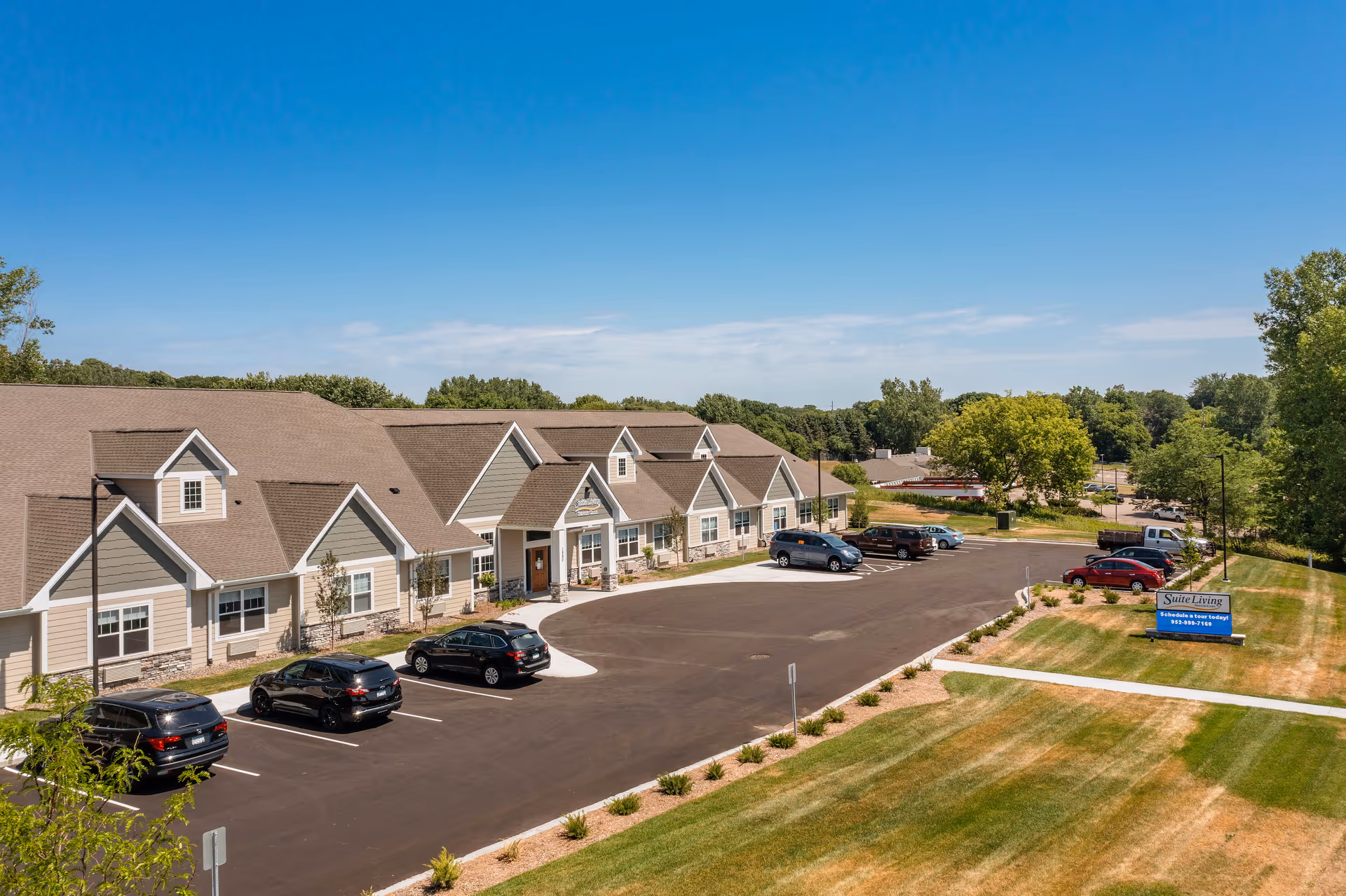 Exterior view of a single-story senior living facility with a parking lot, landscaped lawn, and a sign reading 'Suite Living' under a clear blue sky.