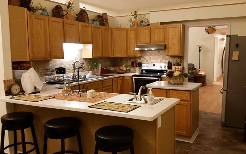 Open-plan kitchen with wooden cabinets, a central island with sink and three bar stools, and stainless steel appliances.