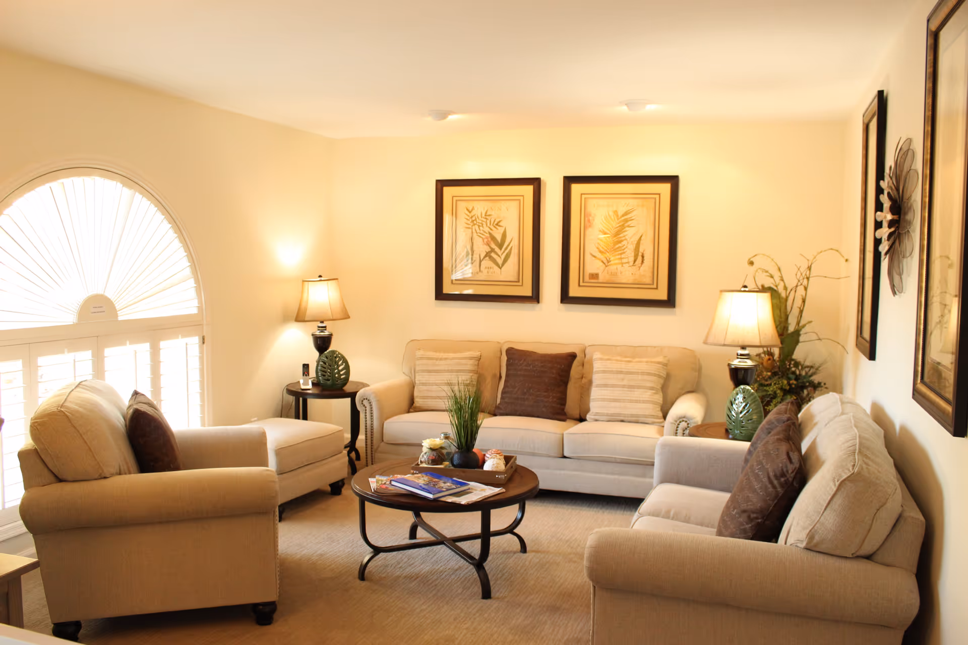 Cozy living room with beige sofas and armchairs around a round coffee table, lamps, framed artwork, and an arched window with shutters.
