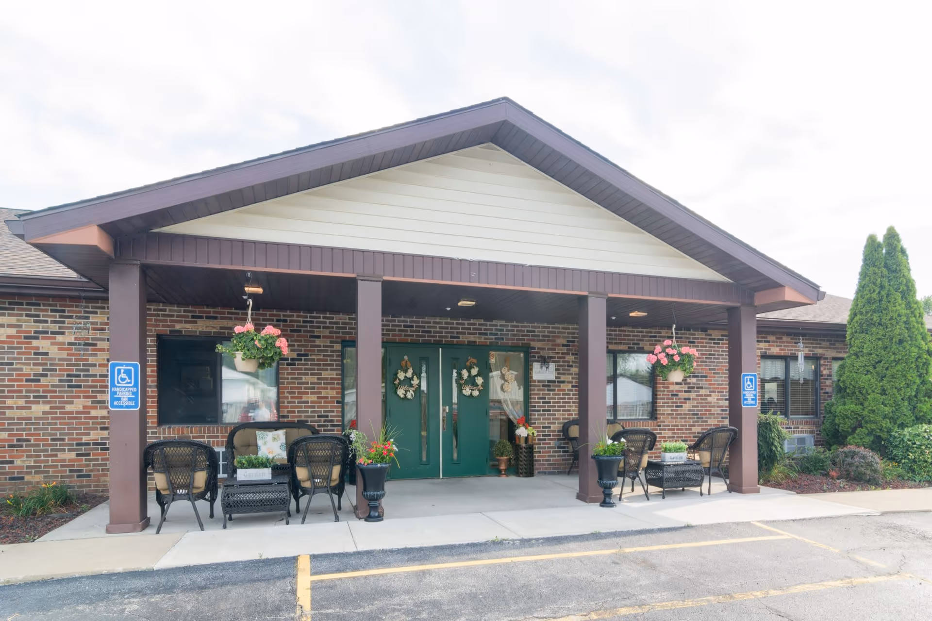 Brick senior living facility entrance with green doors, hanging planters, and outdoor seating.