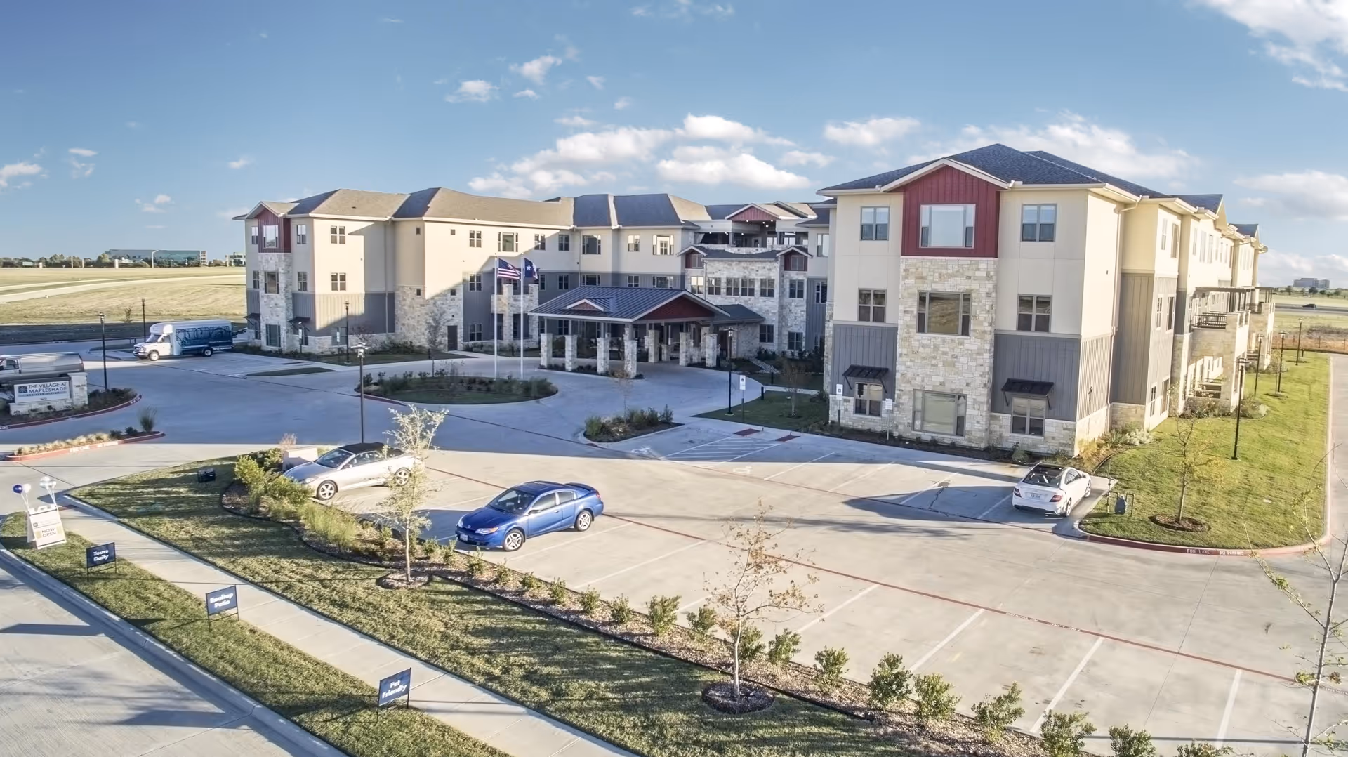 Exterior view of Village on the Park Plano, a multi-story senior living facility with stone and beige siding, surrounded by a parking lot with several cars and landscaped greenery under a blue sky with scattered clouds.