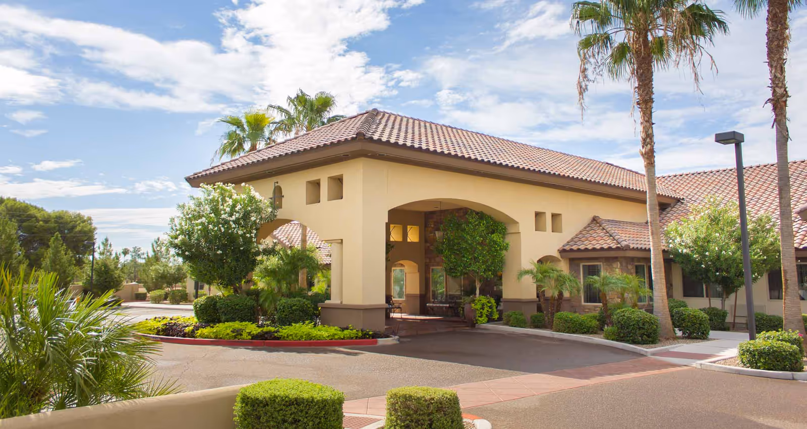 Sunlit senior living facility entrance with covered driveway and tropical landscaping.