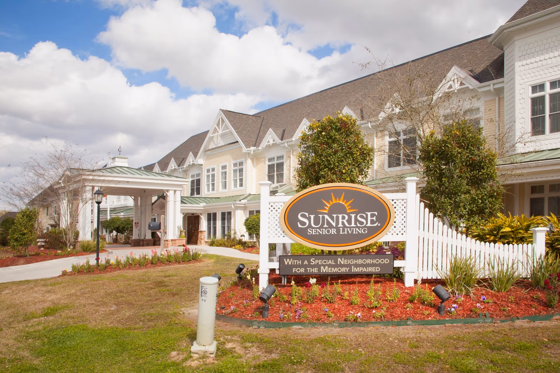 Exterior view of Sunrise Senior Living facility with a large sign in front that reads 'Sunrise Senior Living - With a Special Neighborhood for the Memory Impaired'. The building is a two-story structure with white and beige siding, multiple windows, and a covered entrance. The landscaping includes green grass, bushes, and flowers under a partly cloudy sky.