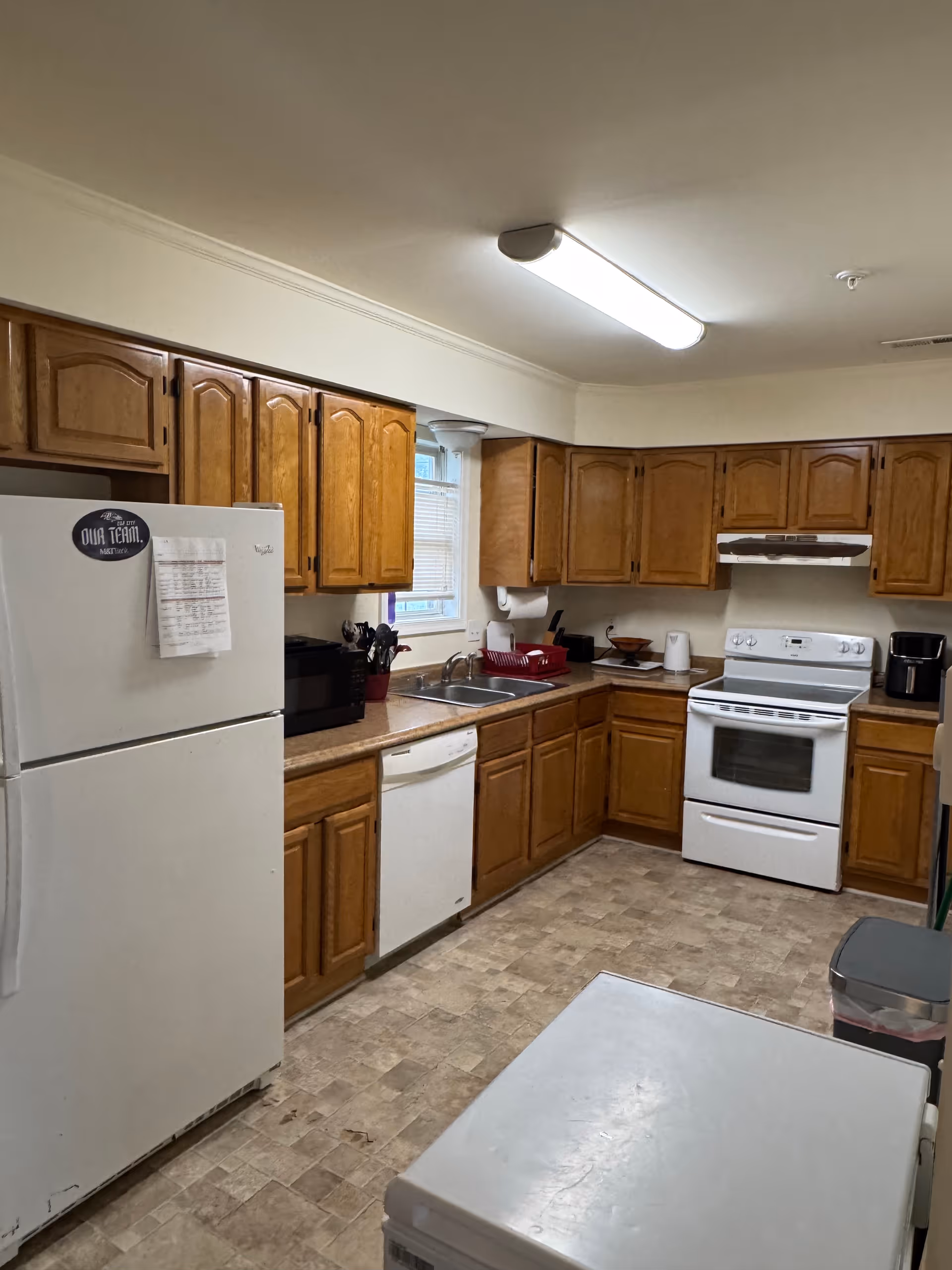 Bright kitchen with wooden cabinets, white appliances, and a double sink under fluorescent lighting.