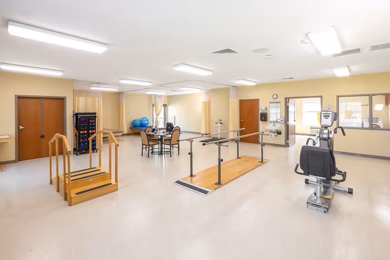 A spacious rehabilitation or physical therapy room with parallel bars for walking practice, a small set of wooden stairs with handrails, exercise equipment including a recumbent bike, a rack of colorful weights, and a table with chairs near windows with yellow curtains.