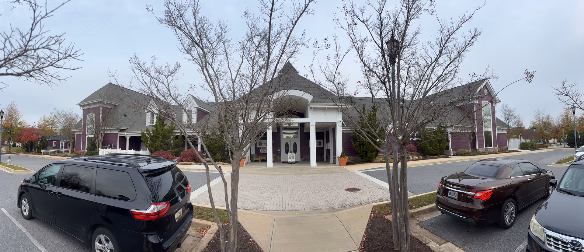 Front exterior view of a senior living facility named Central Parke at Victoria Falls, showing a large building with a peaked roof, white columns at the entrance, and purple siding. There are leafless trees and several parked cars in front of the building under an overcast sky.