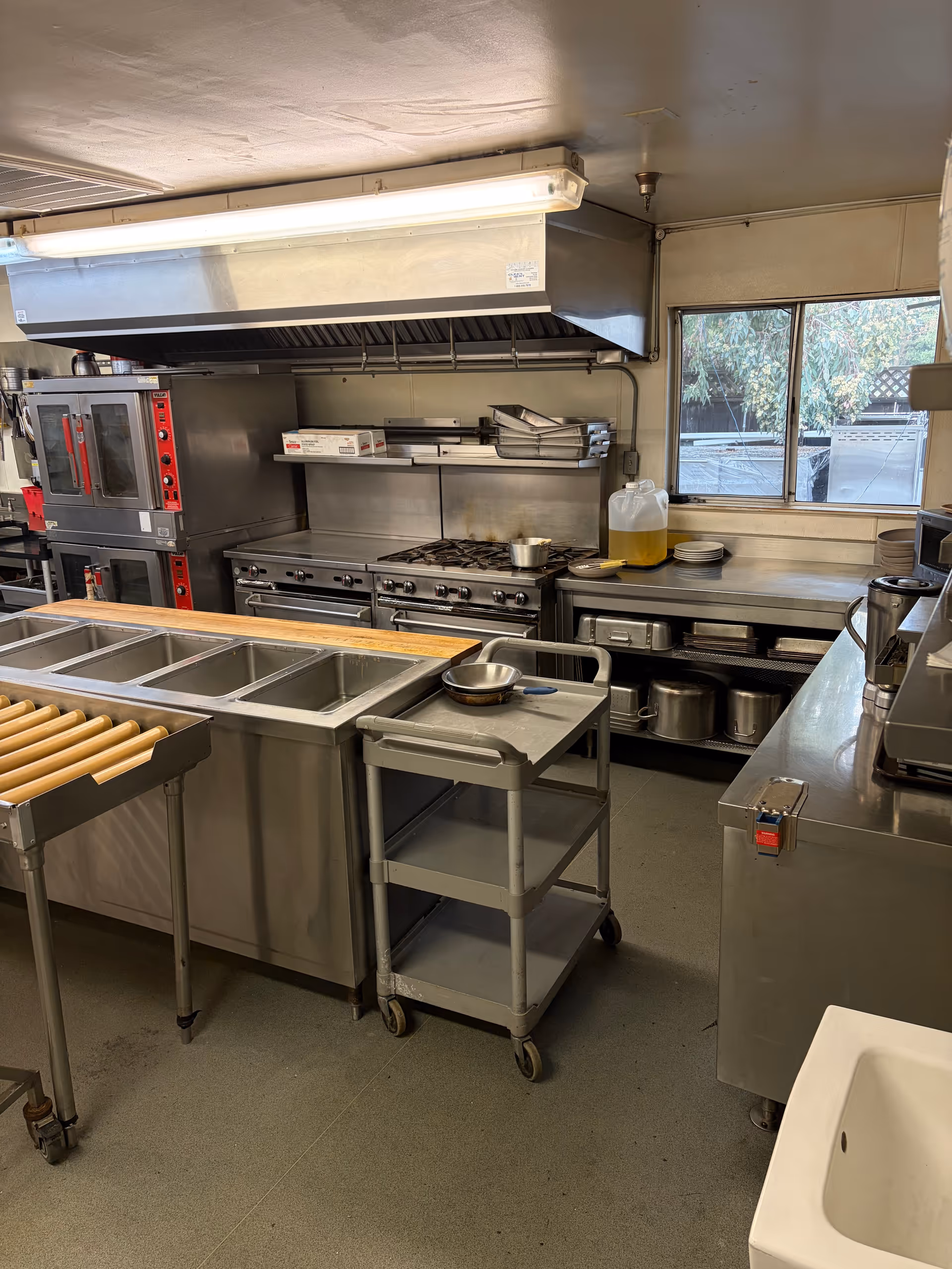 Industrial stainless steel commercial kitchen with ovens, prep tables, and a large vent hood.