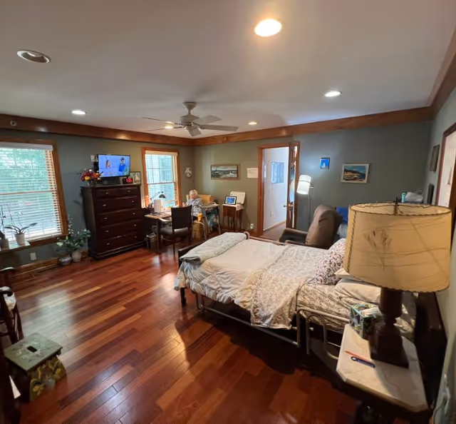 Cozy senior living bedroom with bed, dresser, desk, and seating on polished wooden floors.