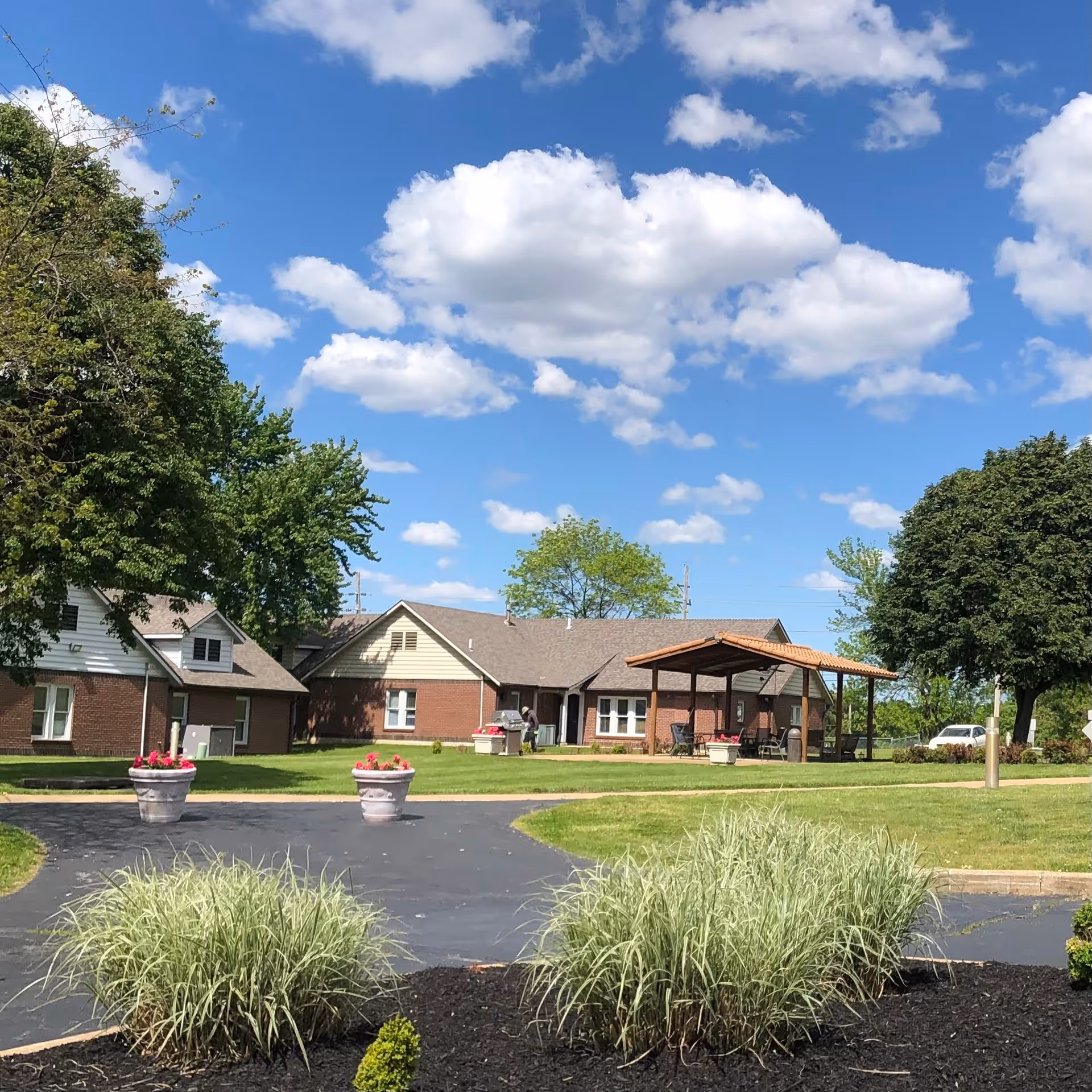 Exterior view of The Gables at Brady Circle- Assisted Living and Memory Care facility showing a single-story brick building with a covered patio area, surrounded by green lawns, trees, and flower pots under a blue sky with scattered clouds.