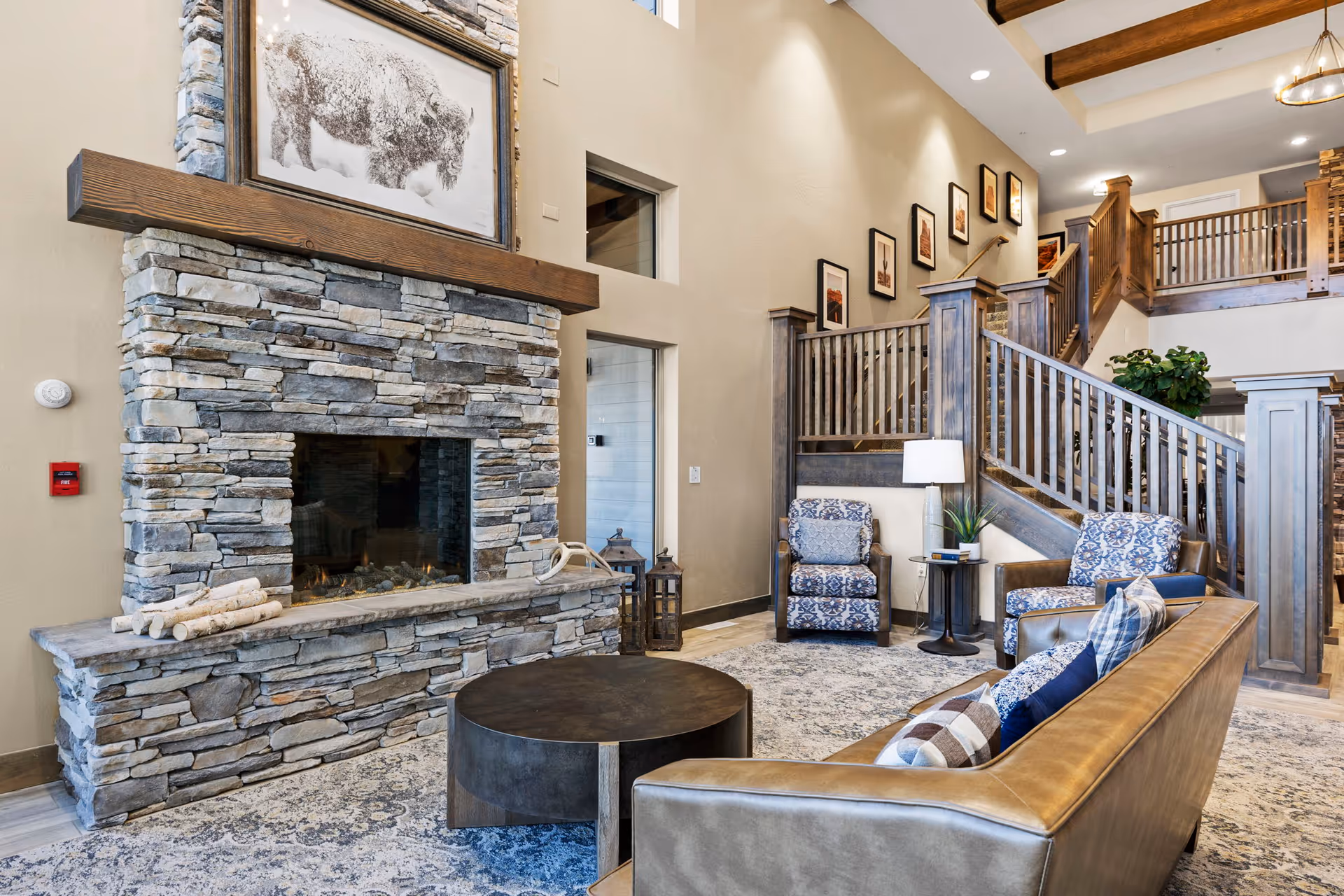 A cozy senior living facility common area featuring a stone fireplace with a wooden mantel and a framed bison artwork above it. The room includes a brown leather sofa with patterned pillows, two patterned armchairs, a round dark coffee table, a side table with a lamp and plant, and a staircase with wooden railings leading to an upper level. The walls are decorated with framed pictures and the space has a warm, inviting atmosphere.