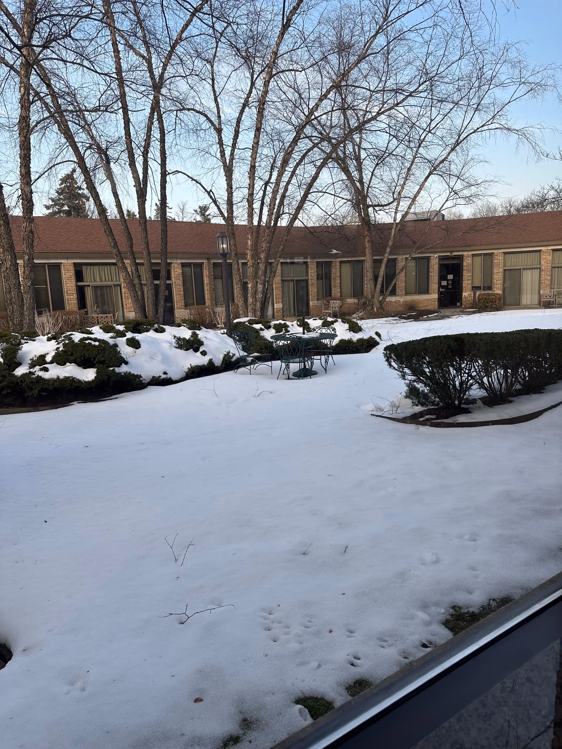 Snow covers a courtyard with leafless trees and a metal table set outside a brick building.