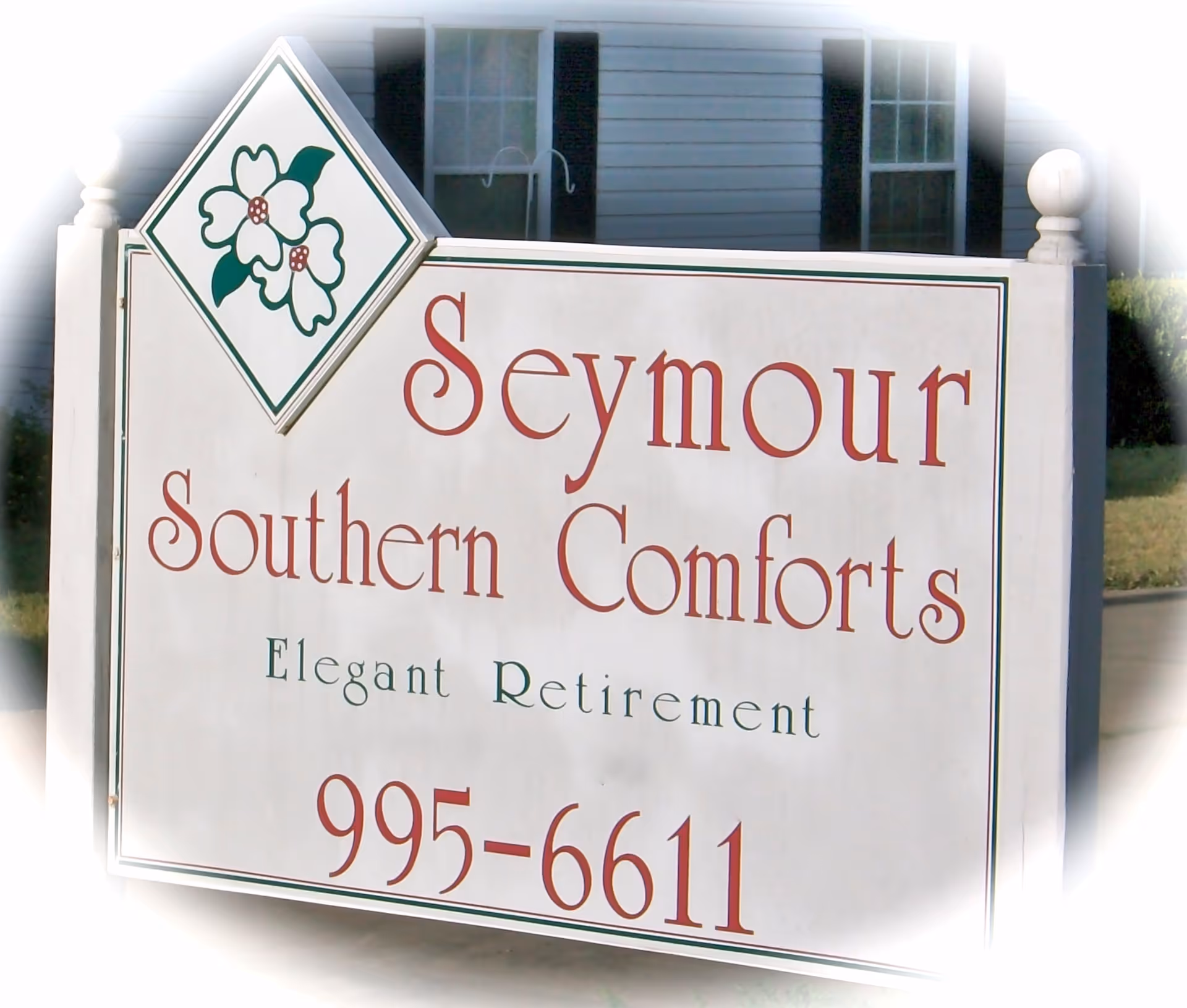 A white outdoor sign with a floral logo at the top left corner that reads 'Seymour Southern Comforts Elegant Retirement' with a phone number 995-6611, placed in front of a building with windows and grass visible in the background.