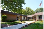 Brick senior living facility front view with walkway, lawn, trees, and an American flag.