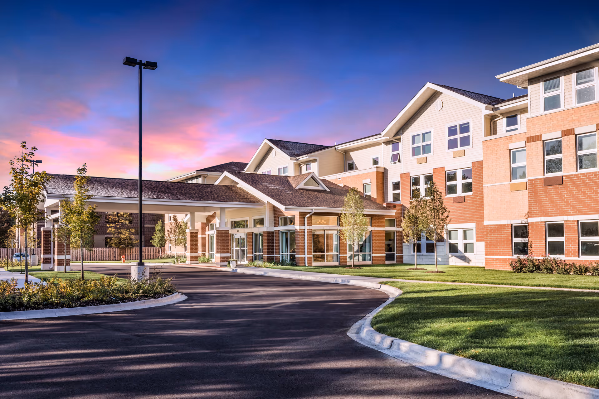 Exterior view of The Sheridan at Tyler Creek senior living facility during sunset, showing a large multi-story building with brick and light-colored siding, a covered entrance, landscaped greenery, and a curved driveway.