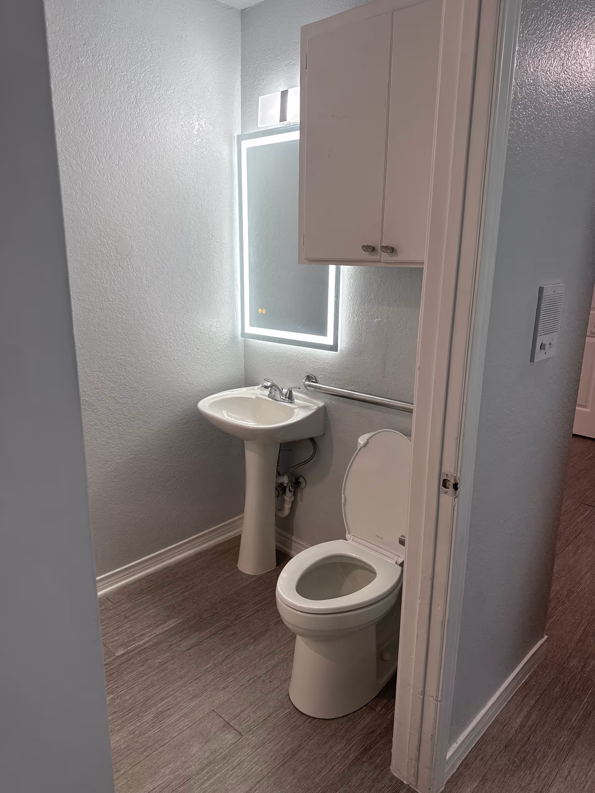 Small bathroom with pedestal sink, mirror, cabinet, toilet, and accessible rail in a facility.