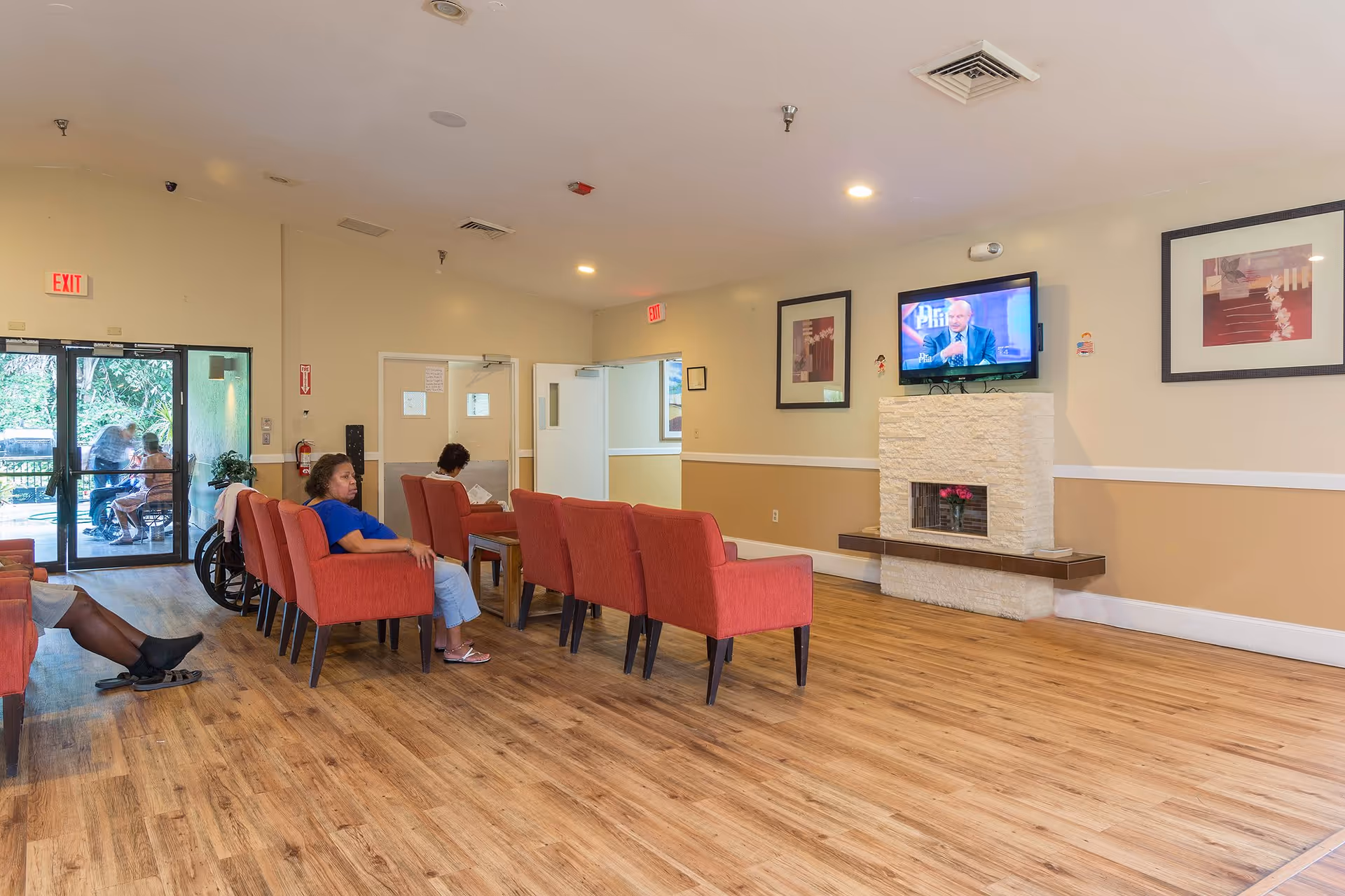 A common area in a senior living facility with several red armchairs arranged in rows facing a wall-mounted television above a decorative fireplace. Two people are seated in the chairs, and there is a glass door leading outside where two more people are visible. The room has wood flooring, beige walls with white trim, and framed artwork on the walls.