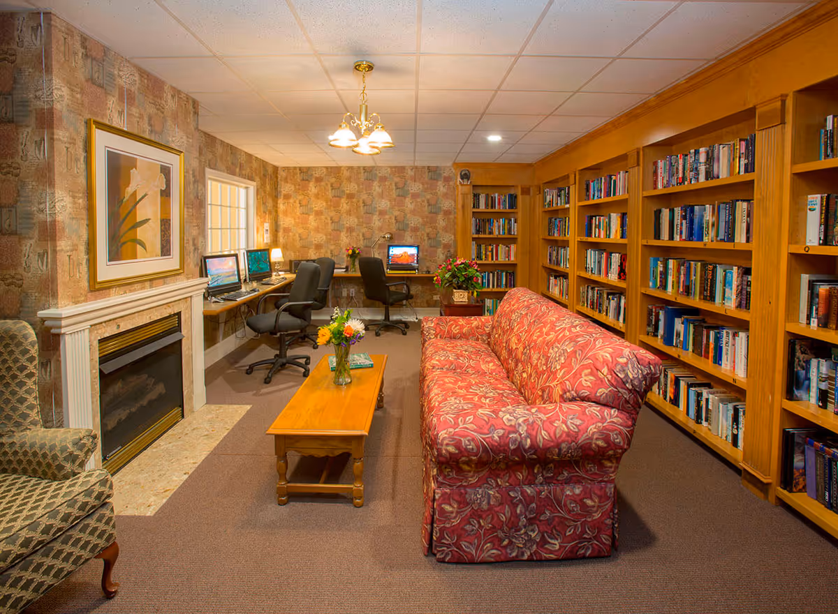 A cozy library and computer room with wooden bookshelves filled with books on the right wall, a red floral patterned couch in the center, a wooden coffee table with a vase of flowers, and a fireplace with a framed floral painting above it on the left. At the back, there are three computer workstations with black office chairs and small lamps, set against a wall with patterned wallpaper.
