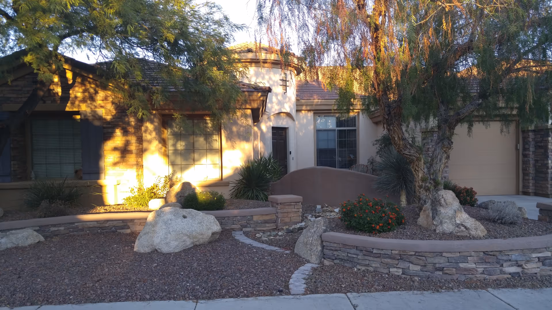 Front exterior view of a single-story assisted living care home with stone and stucco facade, large windows, a garage, desert landscaping including rocks, bushes, and trees, and a curved stone retaining wall.