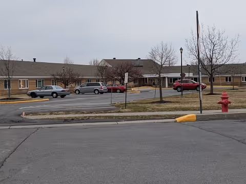Parking lot with several parked cars in front of a single-story brick building with a sloped roof and leafless trees, under an overcast sky.