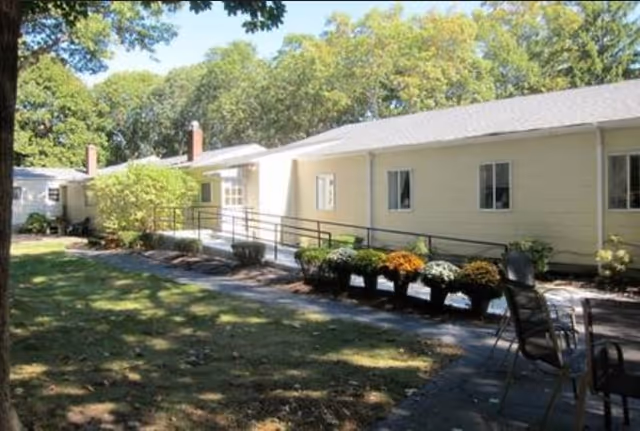 Single-story yellow building with a wheelchair ramp, planters and outdoor seating on a shady lawn.