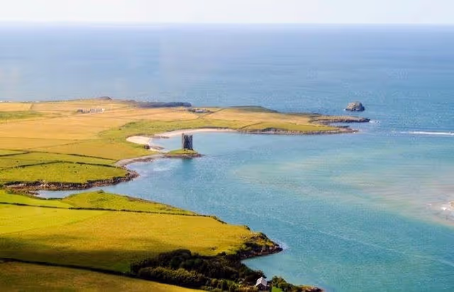 Expansive coastal landscape with green fields meeting a blue sea and rocky shoreline.