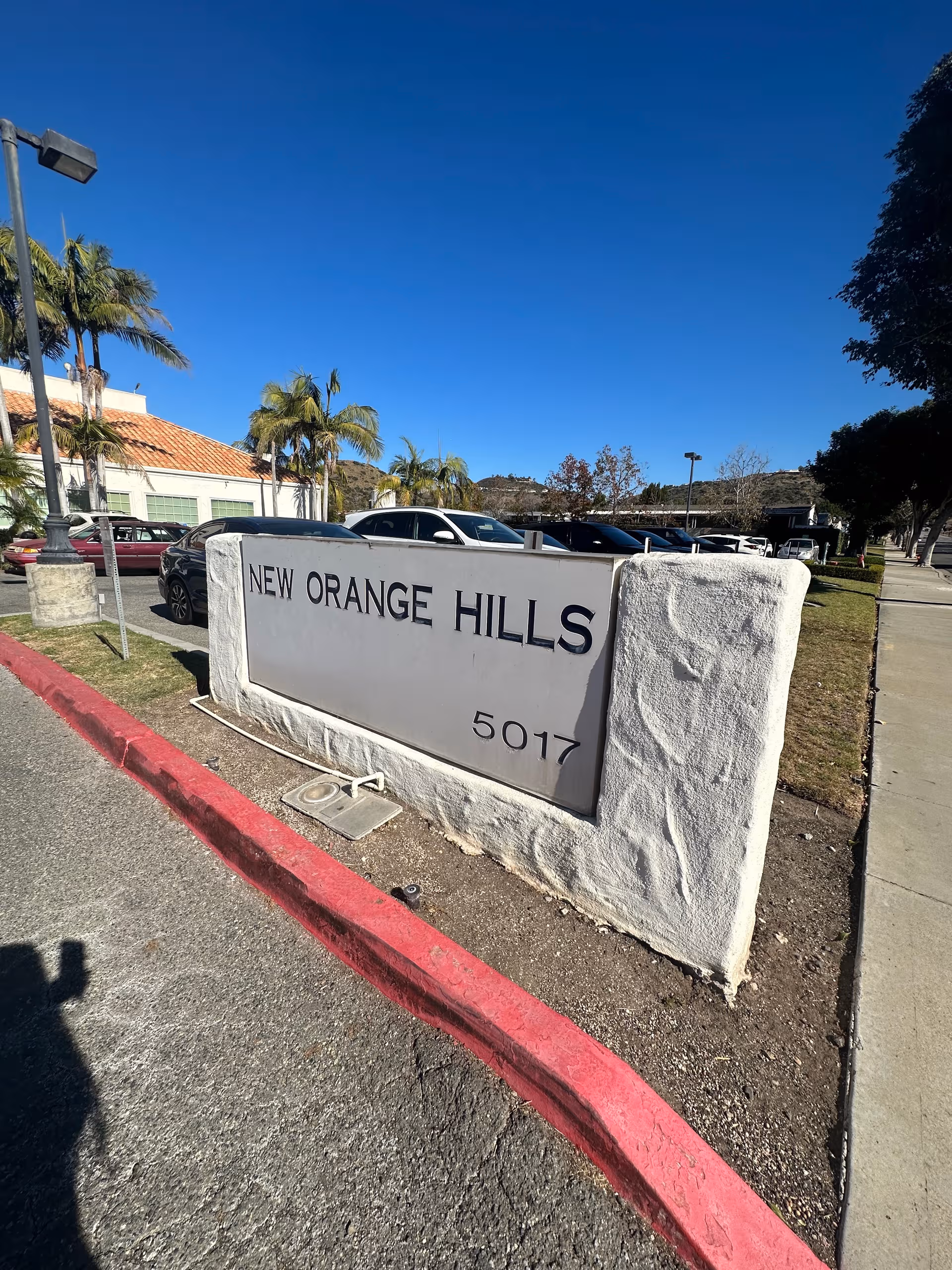 Outdoor view of New Orange Hills entrance sign with parking lot and palm trees.