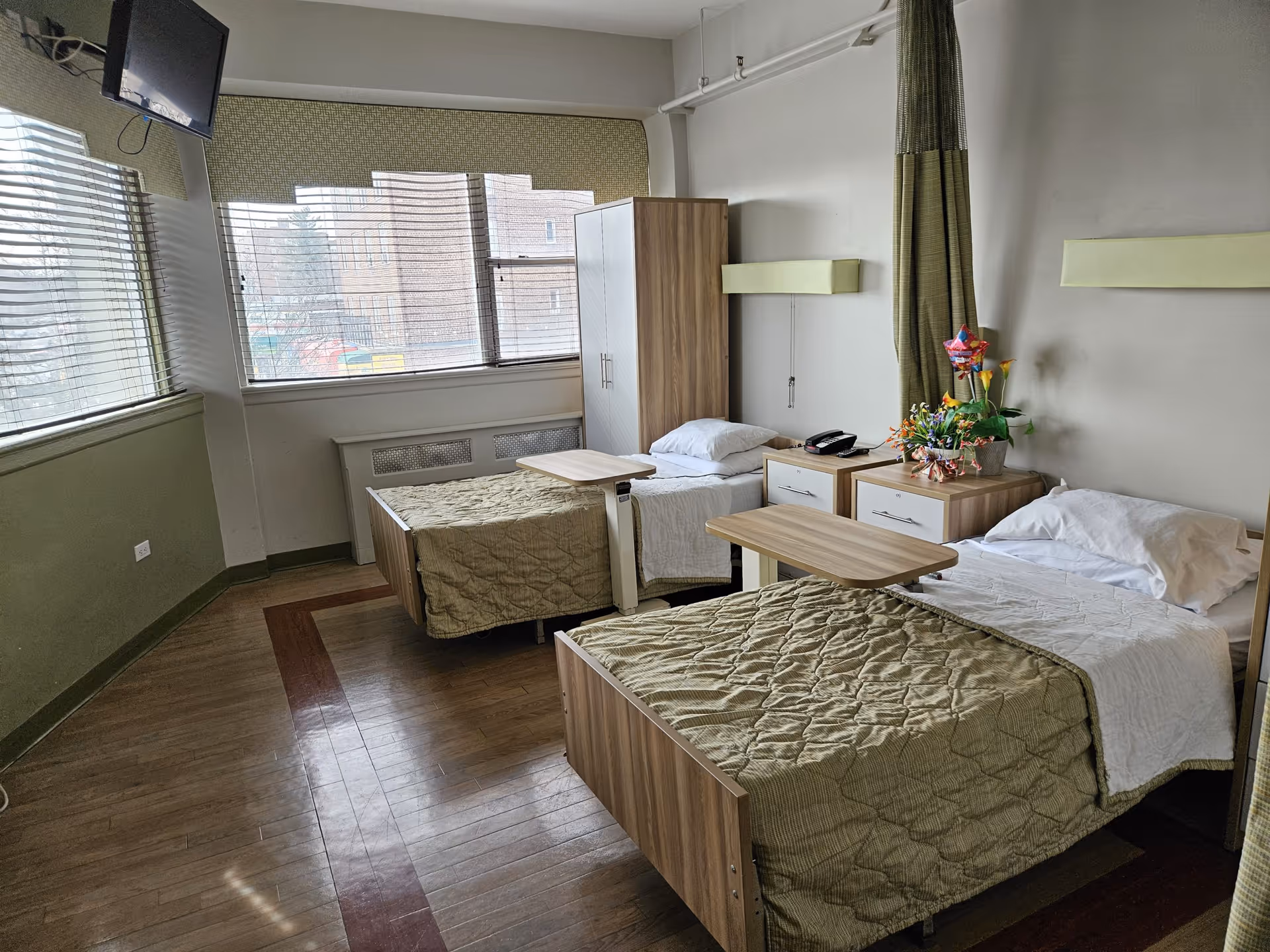 A bright room with two single beds, each with a green quilted blanket and white sheets. Between the beds are two wooden nightstands, one with a telephone and the other with a flower arrangement. There is a wooden wardrobe near the window, which has blinds partially open letting in natural light. A wall-mounted TV is positioned in the corner near the ceiling. The floor is wooden with a darker wood border pattern.