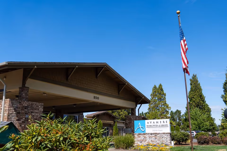 Exterior view of Avamere Rehabilitation of Lebanon building with a covered entrance, brick pillars, green shrubs, an American flag on a flagpole, and a sign displaying the facility name and address under a clear blue sky.