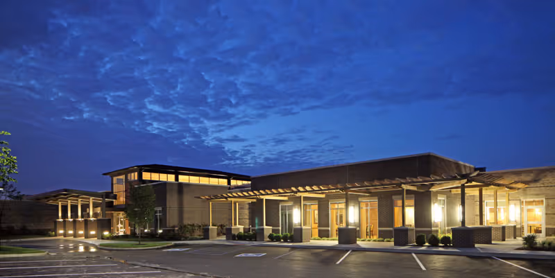 Well-lit senior living facility front entrance at dusk with pergola and parking.