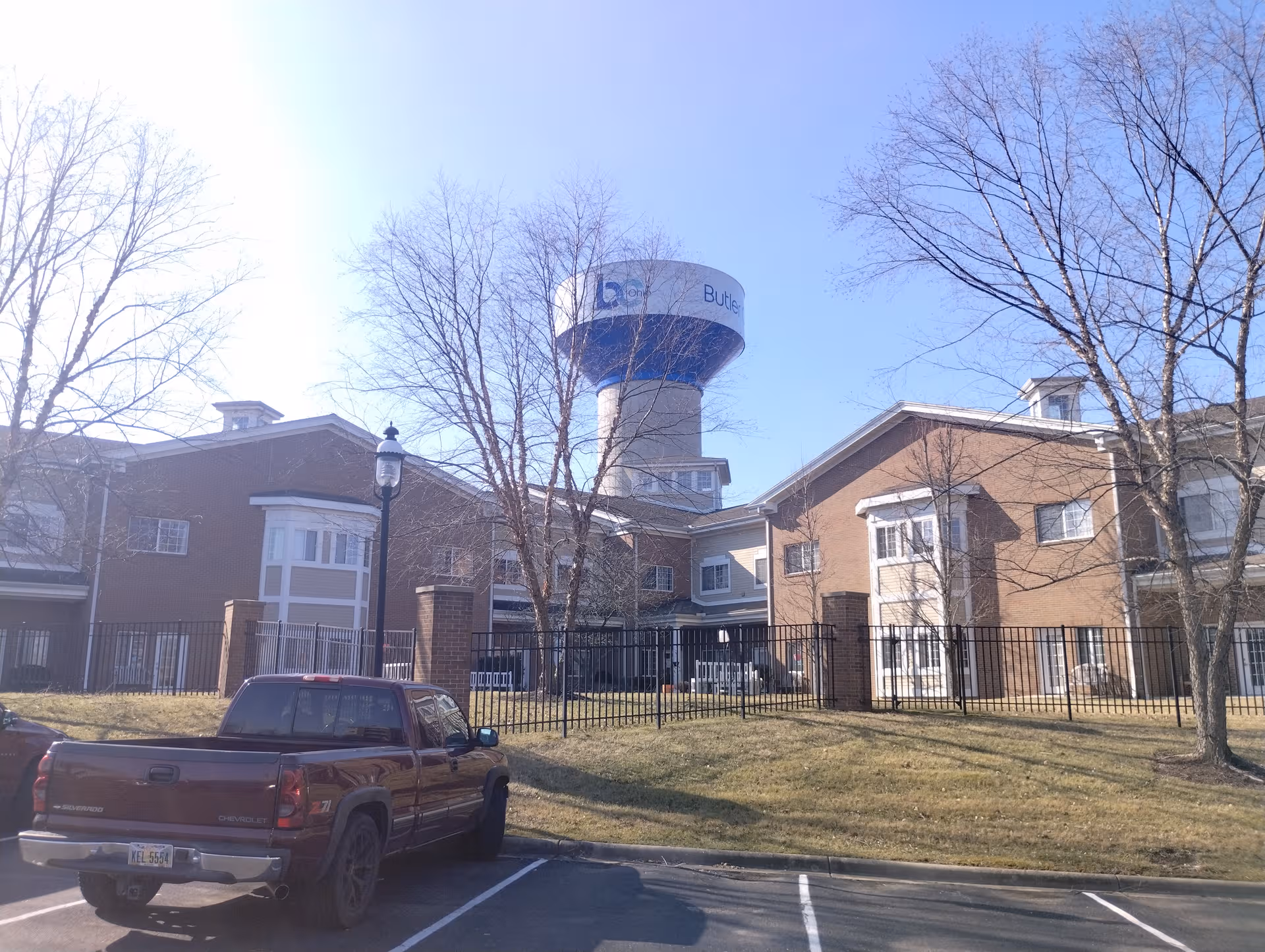 Brick senior living building with a fence, leafless trees, and a parked truck in front.
