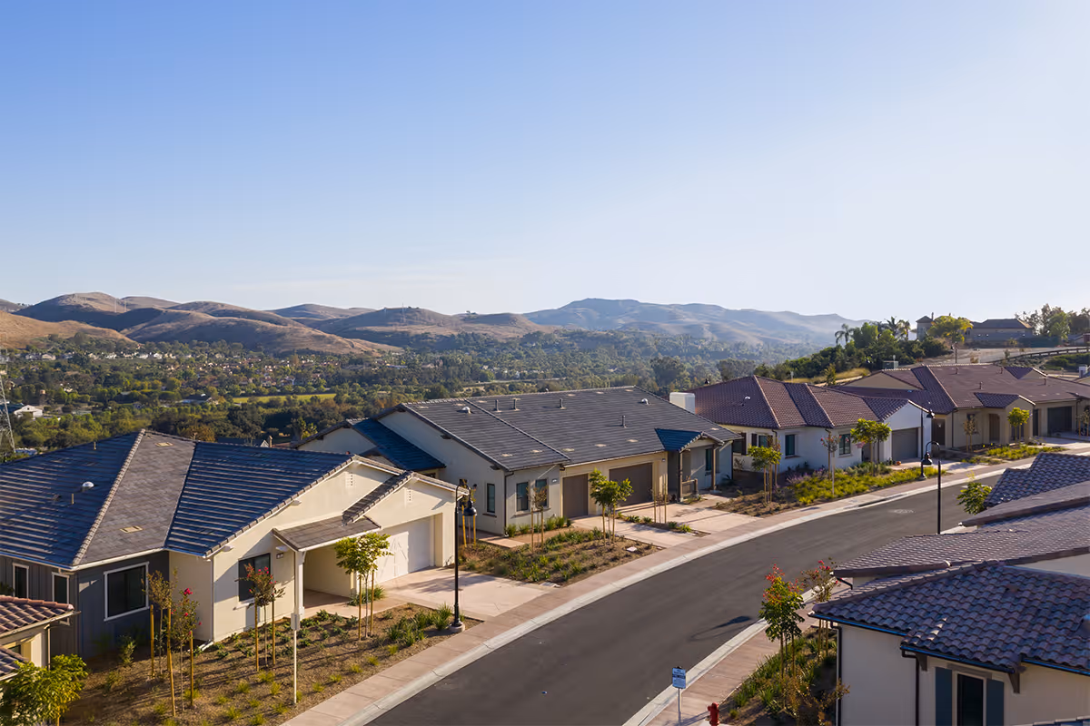 A scenic view of a residential neighborhood with single-story houses featuring tiled roofs, a curved street, young trees, and landscaped yards, set against a backdrop of rolling hills under a clear blue sky.