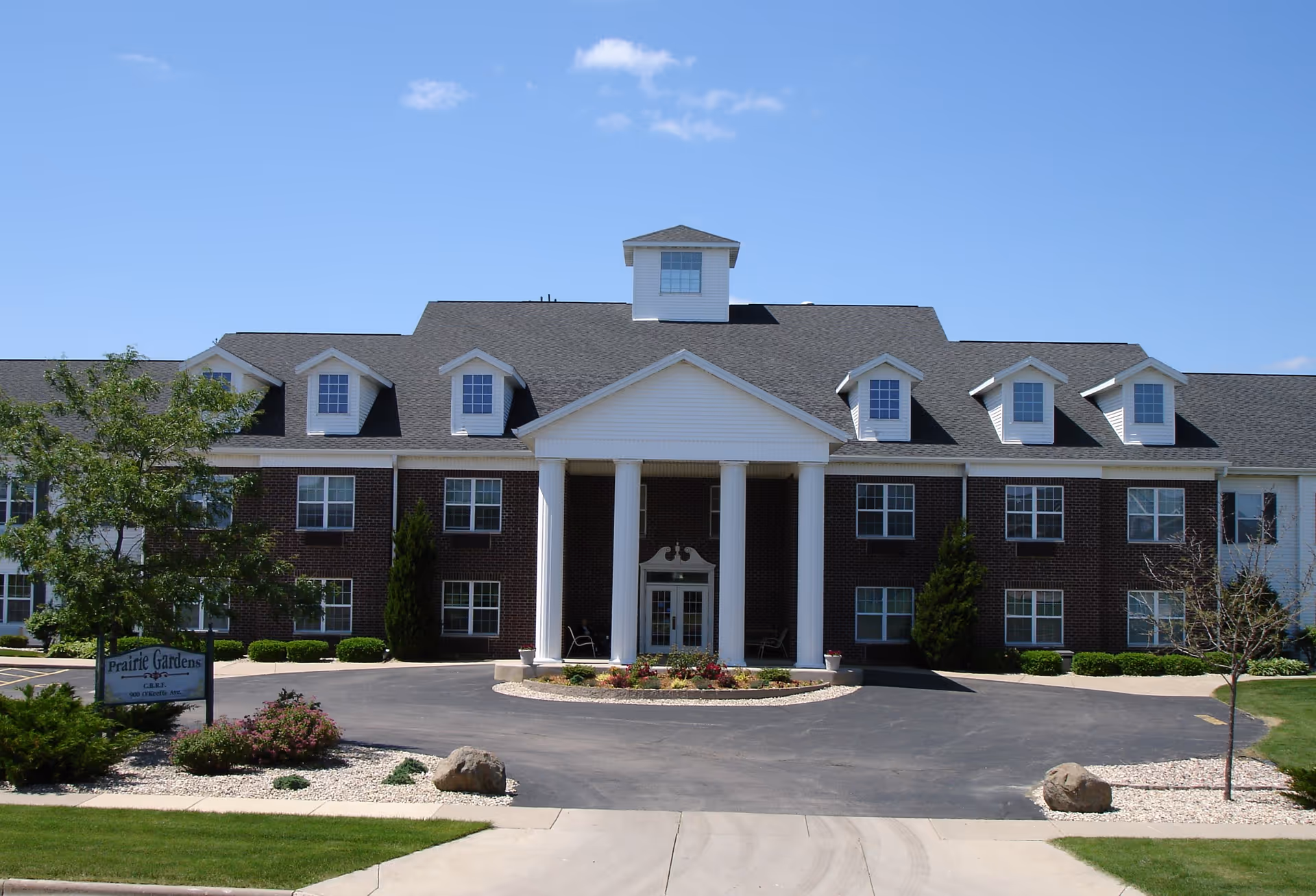 Two-story brick senior living center with a white-columned front portico and landscaped grounds.