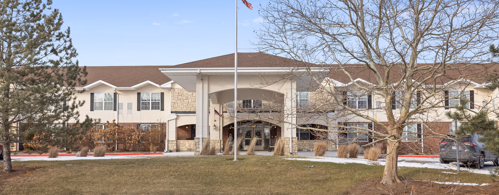 Front exterior view of Bickford of Bourbonnais facility with a covered entrance, an American flag on a flagpole, trees, and a lawn with patches of snow.
