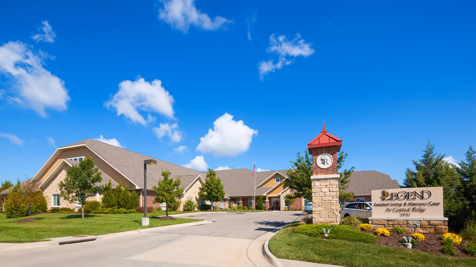 Exterior view of Legend at Capital Ridge, a senior living facility, showing a large building with a peaked roof, surrounded by green lawns, trees, and a clear blue sky with some clouds. There is a stone sign in the foreground with the facility's name and a clock tower next to it.